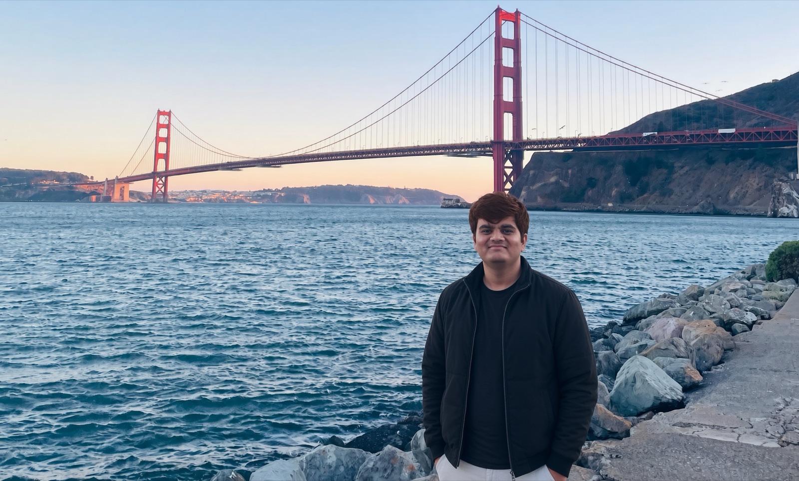 Neel standing on the rocks by the bay at sunset, with the Golden Gate Bridge behind him